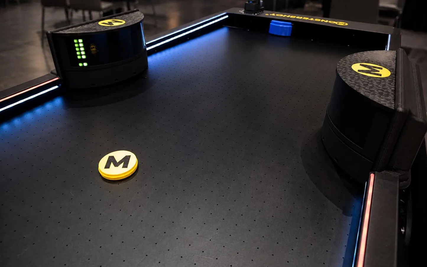 Zwei automatische Bumper mit gelbem M-Logo auf dem beleuchteten Monstershot®-Spielfeld, davor der gelbe Puck