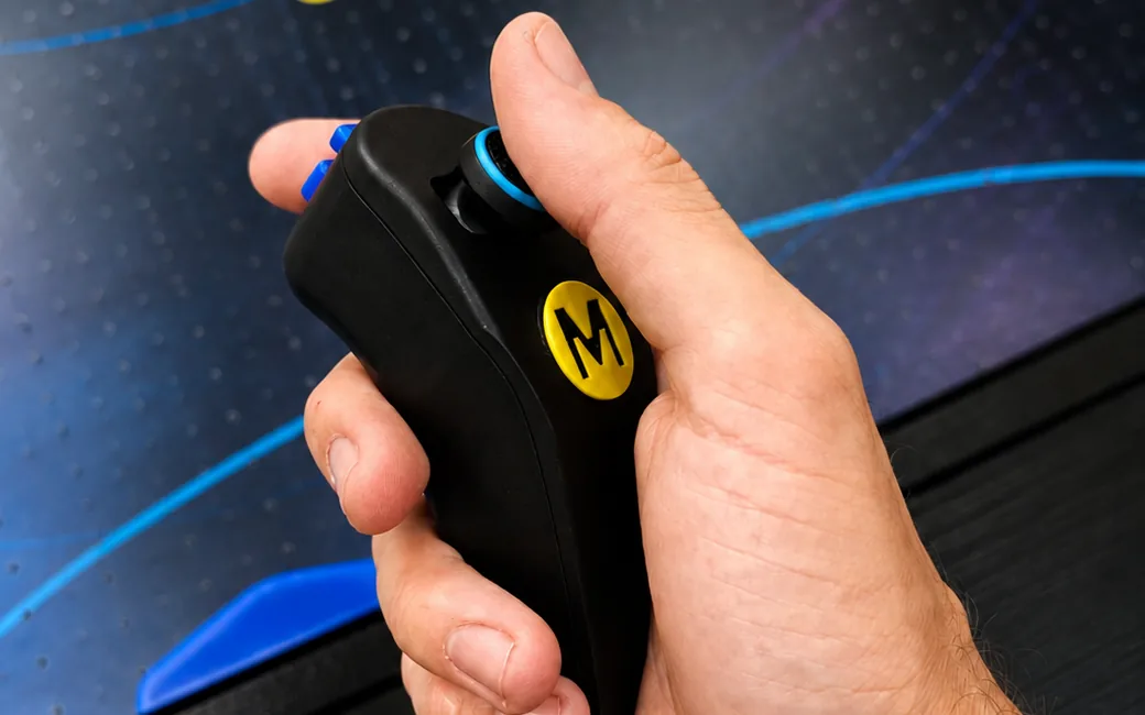 Hand hält den Monstershot®-Handcontroller mit Joystick und gelbem M-Logo über dem Spielfeld